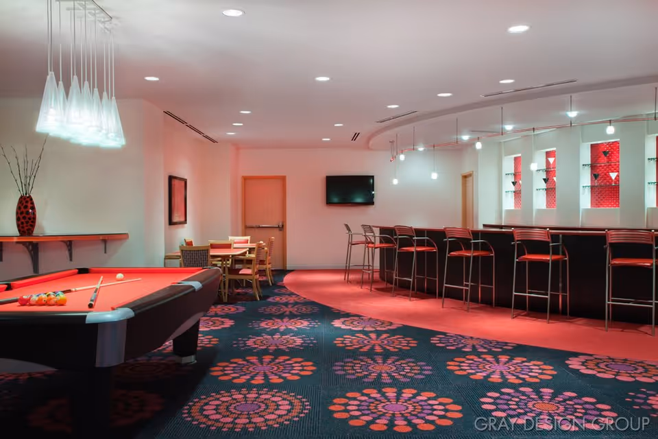 Interior view of a recreational lounge area featuring a red pool table with balls and cue sticks, a seating area with tables and chairs, a bar with high stools, modern pendant lighting, and a wall-mounted TV. The carpet has a colorful floral pattern and the walls are light-colored with red accent lighting behind glass shelves.