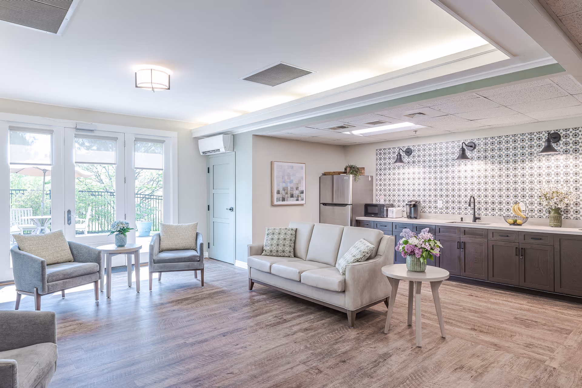 Bright, open common room with a sofa and armchairs facing a small table, a kitchenette with patterned backsplash and cabinets, and glass doors leading to an outdoor patio.