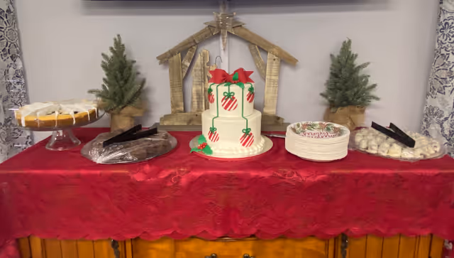 A festive dessert table with a red tablecloth featuring a two-tier white cake decorated with red and green Christmas ornaments and a red bow on top. The table also has a wooden nativity scene backdrop, two small potted evergreen trees, plates with various desserts including a bundt cake, brownies, and white powdered treats, along with stacks of holiday-themed paper plates.