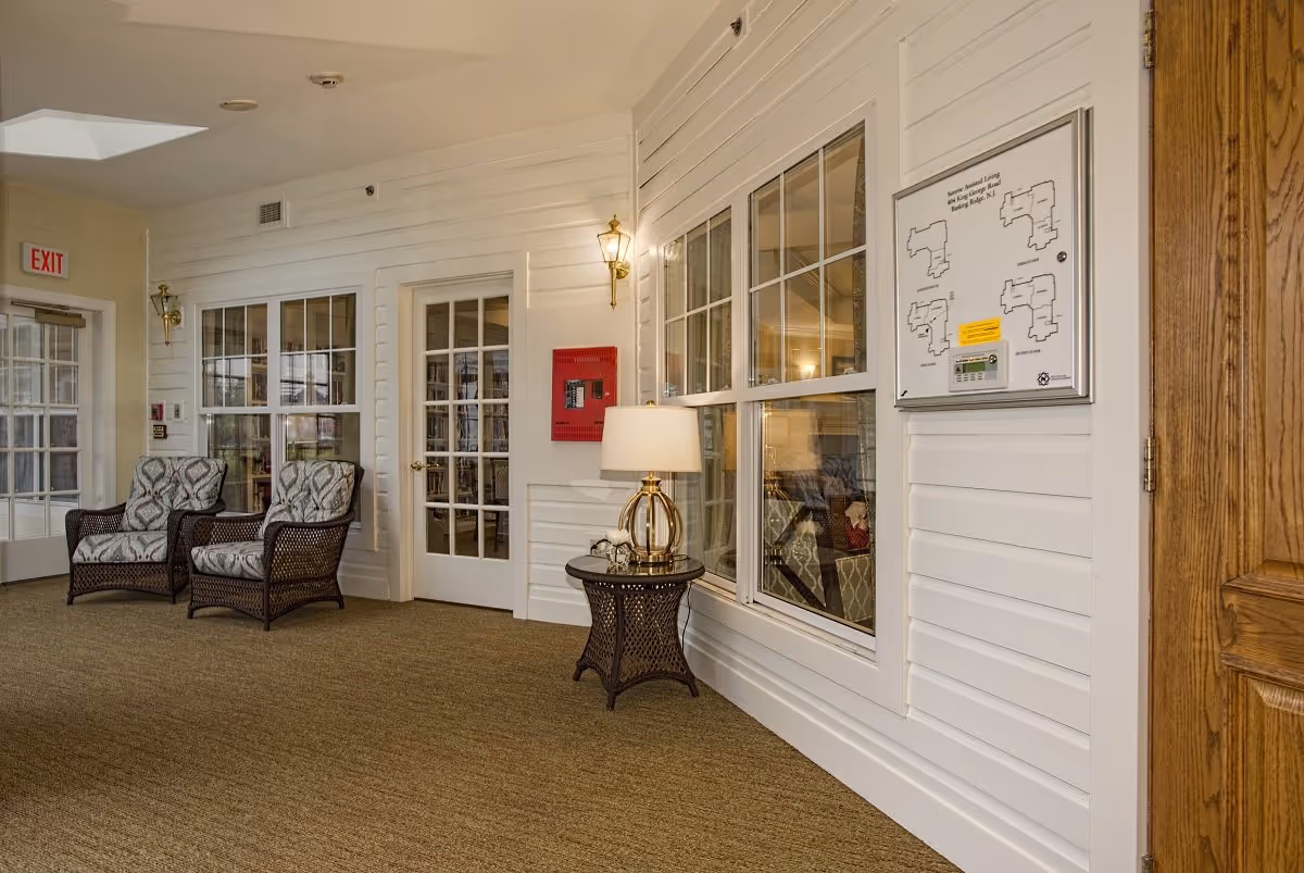 Interior hallway area with two cushioned wicker chairs, a small round wicker table with a lamp, white paneled walls, a fire alarm panel, and a floor plan mounted on the wall. There are glass-paned doors and windows, and an exit sign is visible above a door.