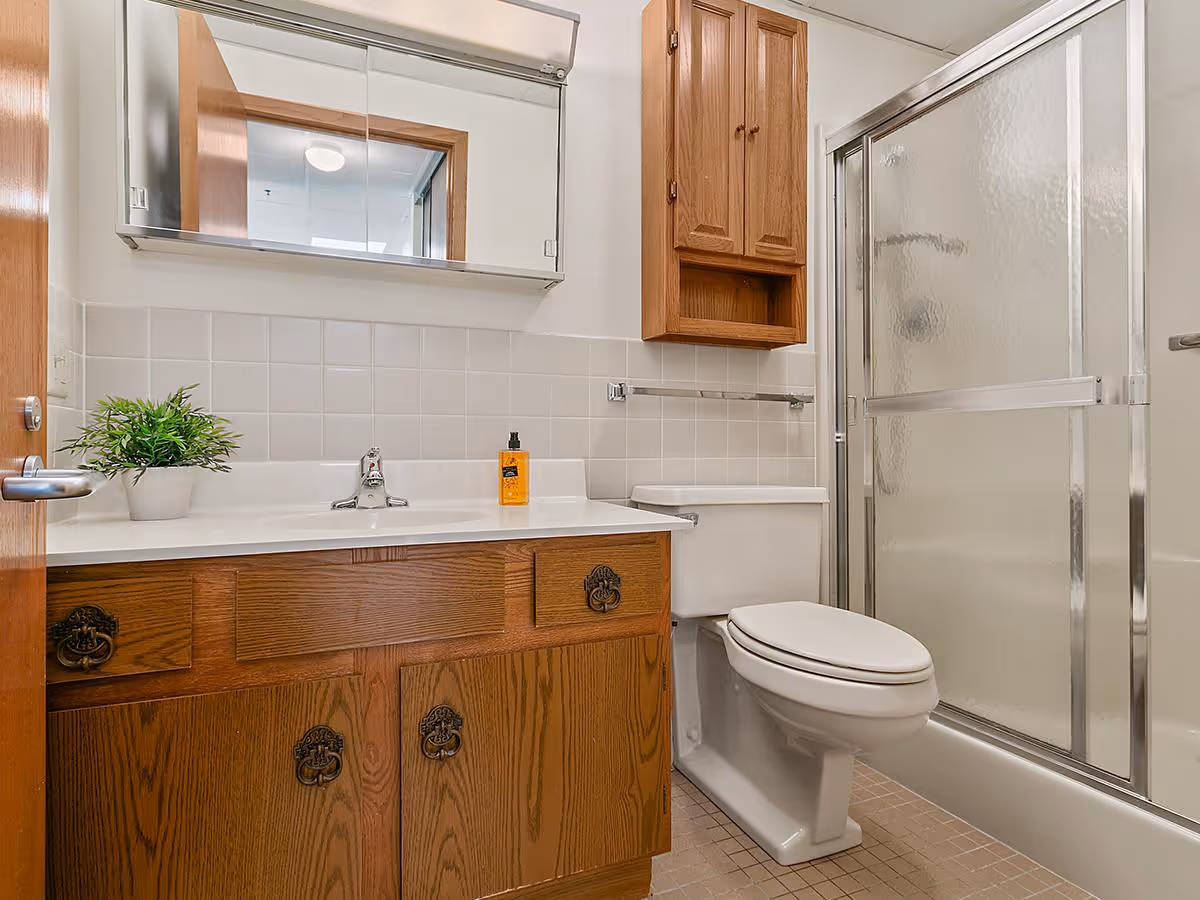 A clean bathroom with a wooden vanity cabinet, a white sink, a small green potted plant, a soap dispenser, a toilet, a wall-mounted wooden cabinet, a towel rack, and a shower with frosted glass sliding doors.