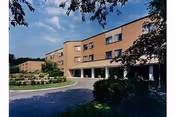 Exterior view of a multi-story brick building with large windows, surrounded by landscaped greenery and a driveway in front under a partly cloudy sky.