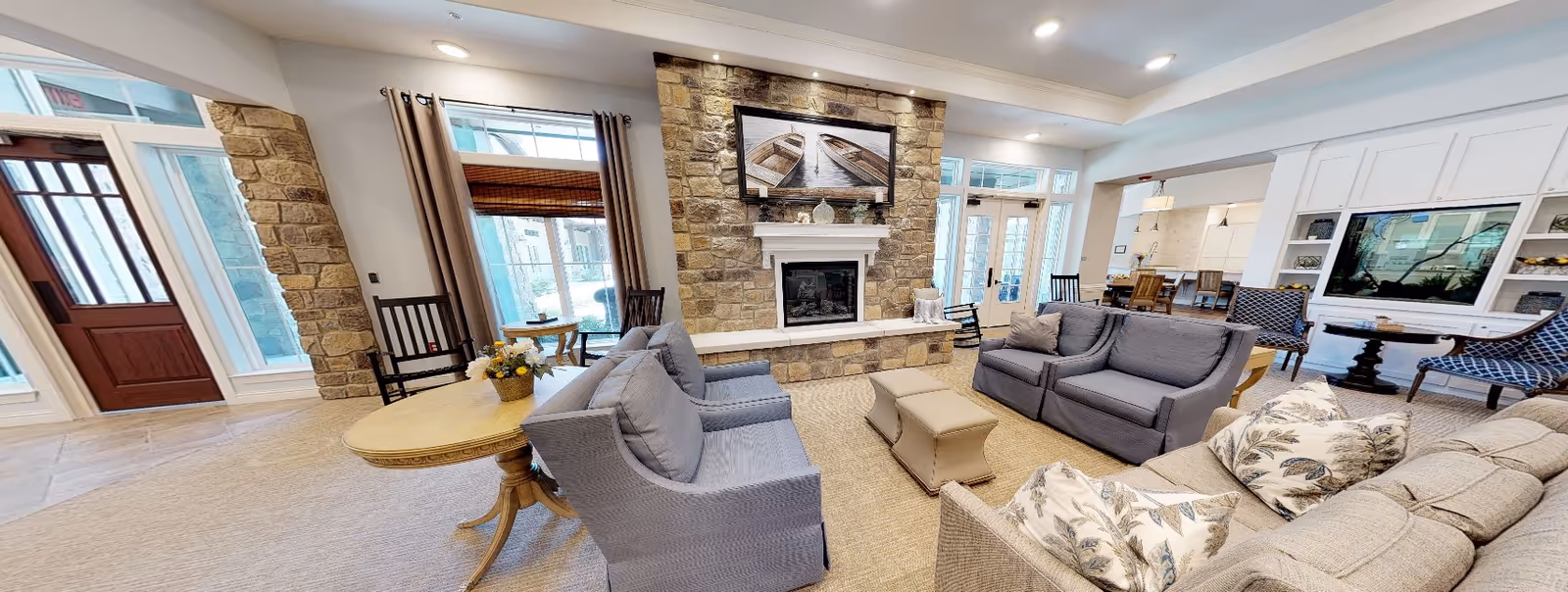 A spacious and well-lit living room area in a senior living facility featuring a stone fireplace with a framed picture of two boats above it. The room has multiple seating options including sofas, armchairs, and wooden rocking chairs arranged around the fireplace. Large windows with curtains and a glass door allow natural light to enter. The room also includes a built-in white entertainment center with a large TV and shelves, and a dining area is visible in the background.
