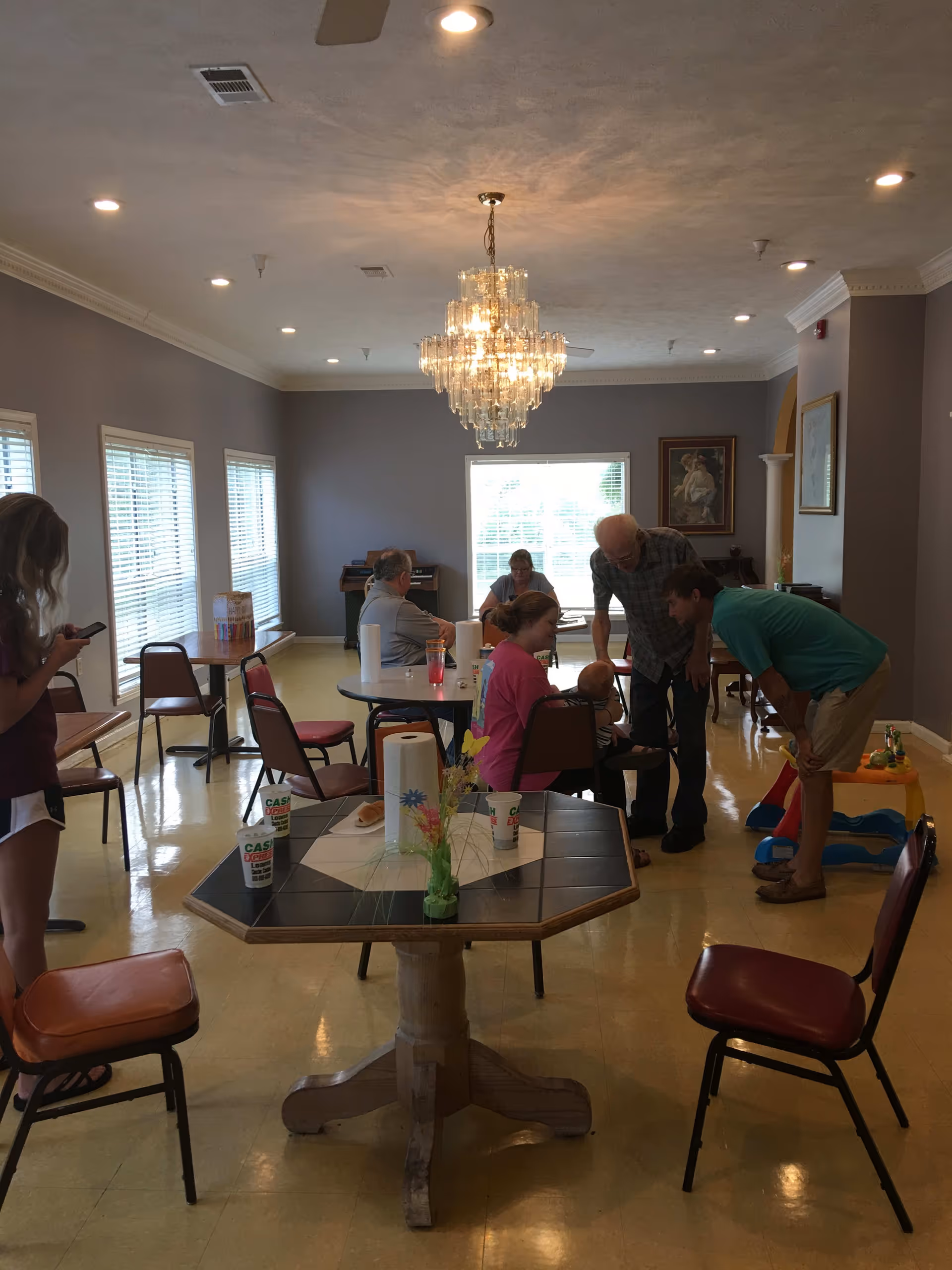 A common area in an assisted living facility with several people interacting. There are tables and chairs arranged around the room, a chandelier hanging from the ceiling, and large windows letting in natural light. Some people are seated while others are standing and engaging with a child. The walls are painted gray and decorated with framed artwork.