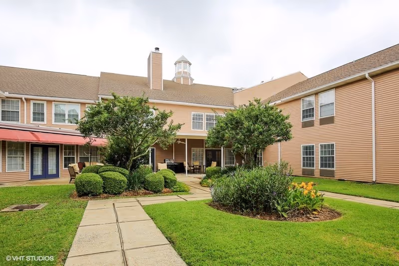 Outdoor courtyard area of a senior living facility with well-maintained green grass, shrubs, and trees. The building surrounding the courtyard is two stories with beige siding, multiple windows, and a covered patio area with chairs and tables. The sky is overcast.