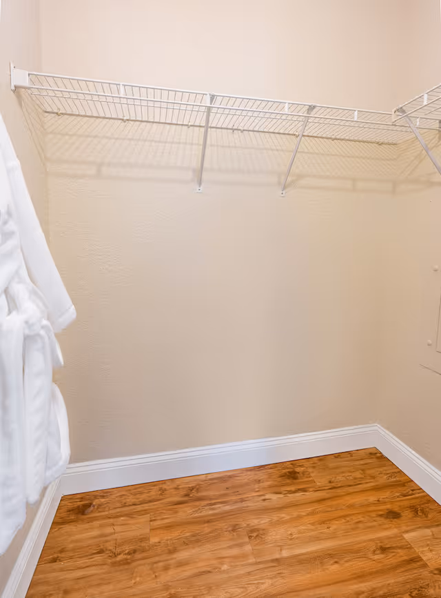 Empty closet with beige textured walls, a wooden floor, and a white wire shelf mounted on the wall. Two white robes are hanging on the left side.
