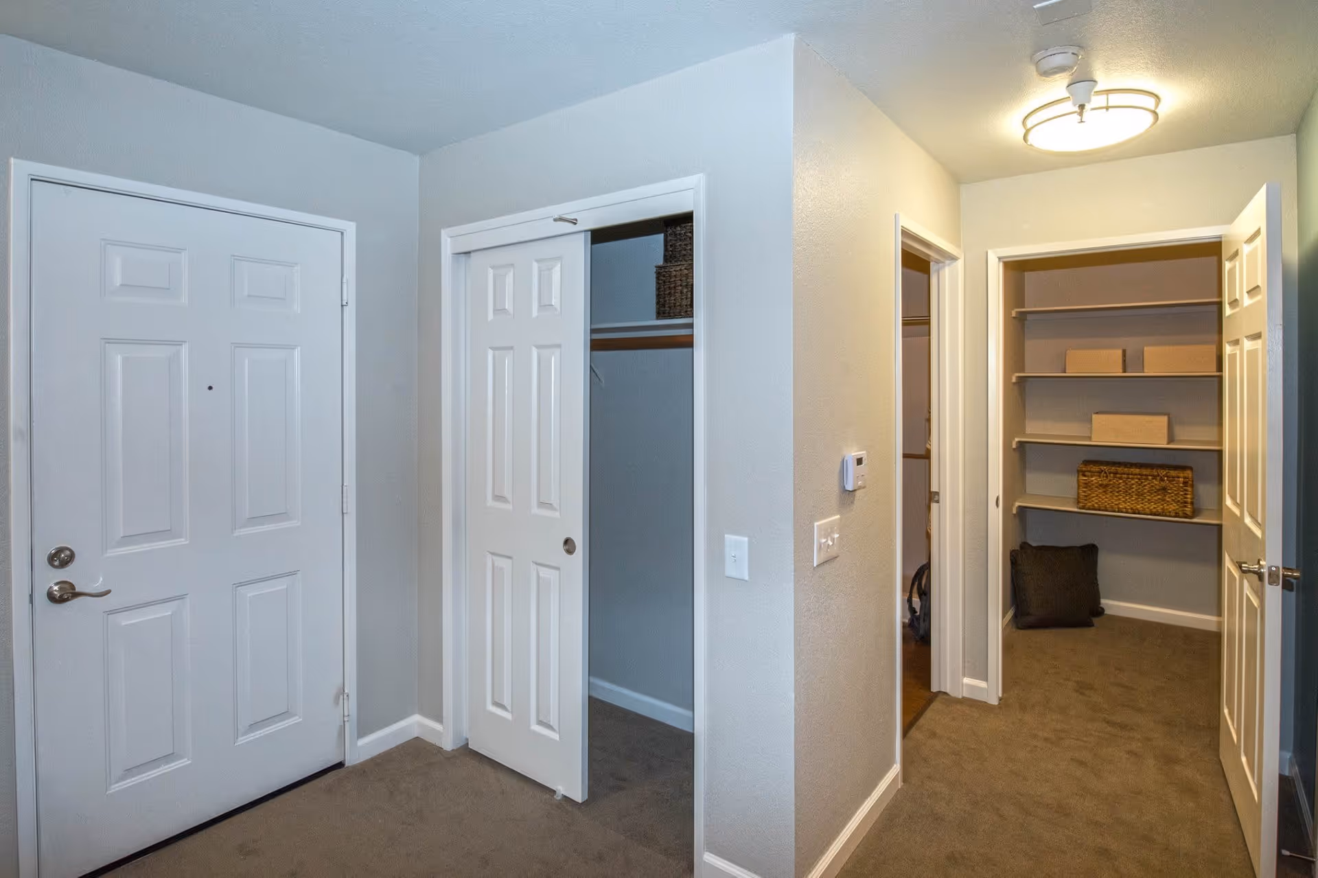 Interior view of a hallway in a senior living facility with a closed white entrance door, an open closet with sliding doors, and an open door leading to a small room with shelves holding storage boxes and a basket. The walls are light-colored and the floor is carpeted.