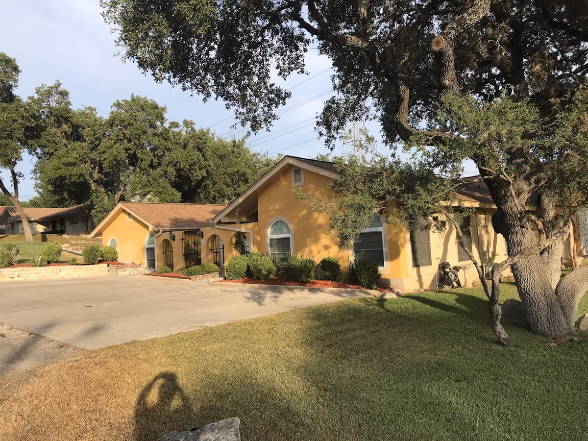 Front view of a single-story yellow residential building with arched windows, a driveway, lawn, and large trees.
