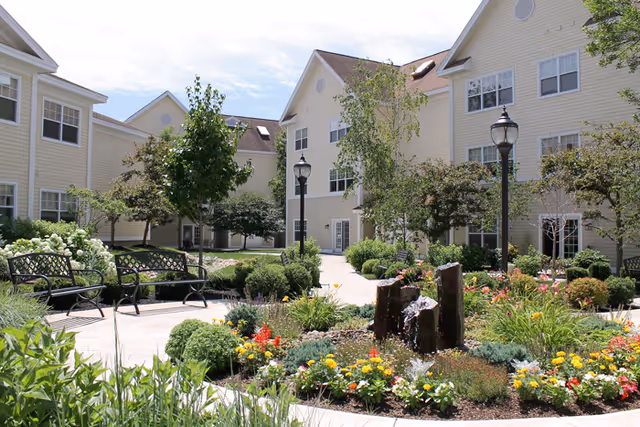 Sunny landscaped courtyard with benches, colorful flower beds, a small fountain, and multi-story cream-colored residential buildings.