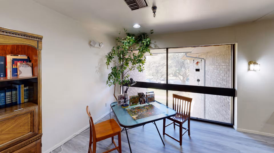A small room with a glass table and two wooden chairs. On the table, there is a puzzle in progress, a desk lamp, and some books. A tall green plant is placed near the corner by a large window that lets in natural light. A wooden bookshelf filled with books is visible on the left side. The floor is light-colored wood, and the walls are white with a wall-mounted light fixture on the right.