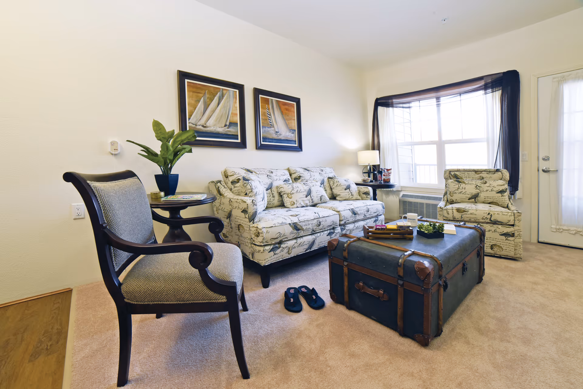 A cozy living room with a patterned sofa and matching armchair, a wooden armchair with cushioned seat, a dark wooden side table with a potted plant, a vintage trunk used as a coffee table, and a large window with sheer curtains letting in natural light.