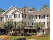 Exterior view of a multi-story residential building with beige siding, green shutters, and a peaked roof. There are several trees and well-maintained bushes in front of the building, along with a flagpole displaying the American flag. The sky is clear and blue.