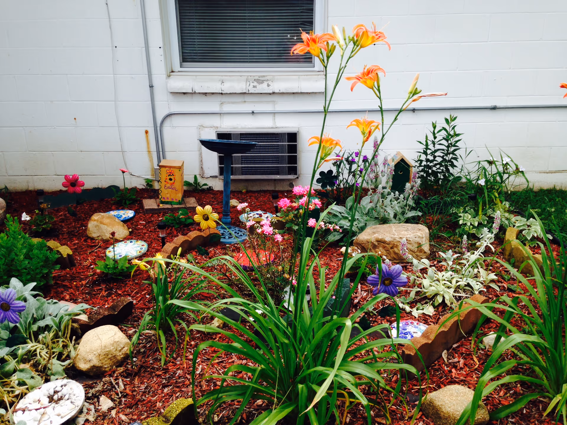A colorful garden bed with various flowers, plants, decorative stones, and garden ornaments in front of a white building wall with a window and an air conditioning unit.