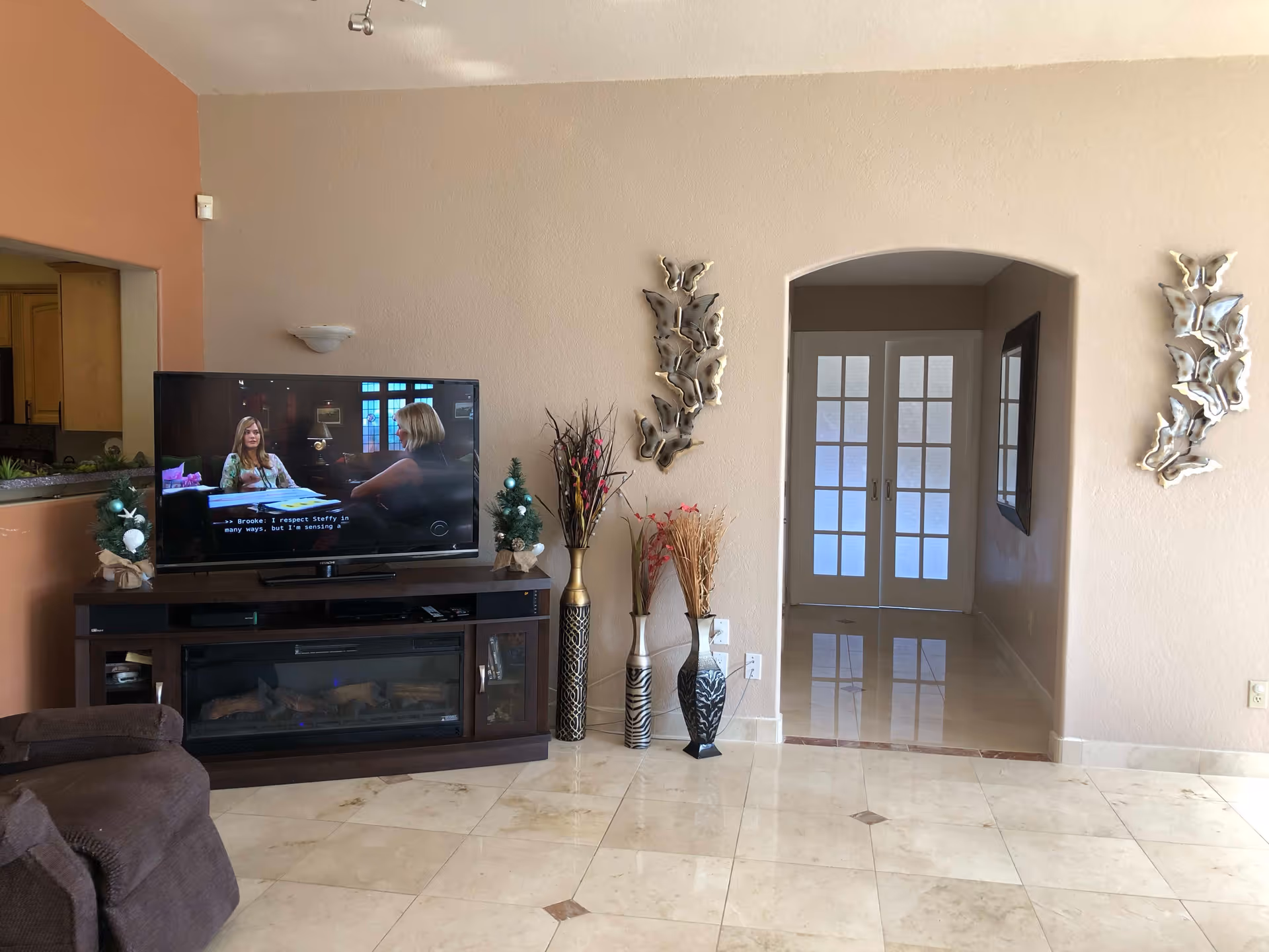 A living room with a large flat-screen TV on a dark wooden stand that also houses an electric fireplace. Two small decorated Christmas trees are placed on either side of the TV. The room has beige walls with two metallic butterfly wall decorations on either side of an arched doorway leading to a hallway with double glass doors. There are three decorative vases with dried plants next to the TV stand. The floor is tiled with light-colored tiles and a brown recliner chair is partially visible on the left.