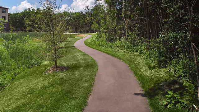 A paved walking path winding through green grass and trees with a building visible in the distance under a partly cloudy sky.