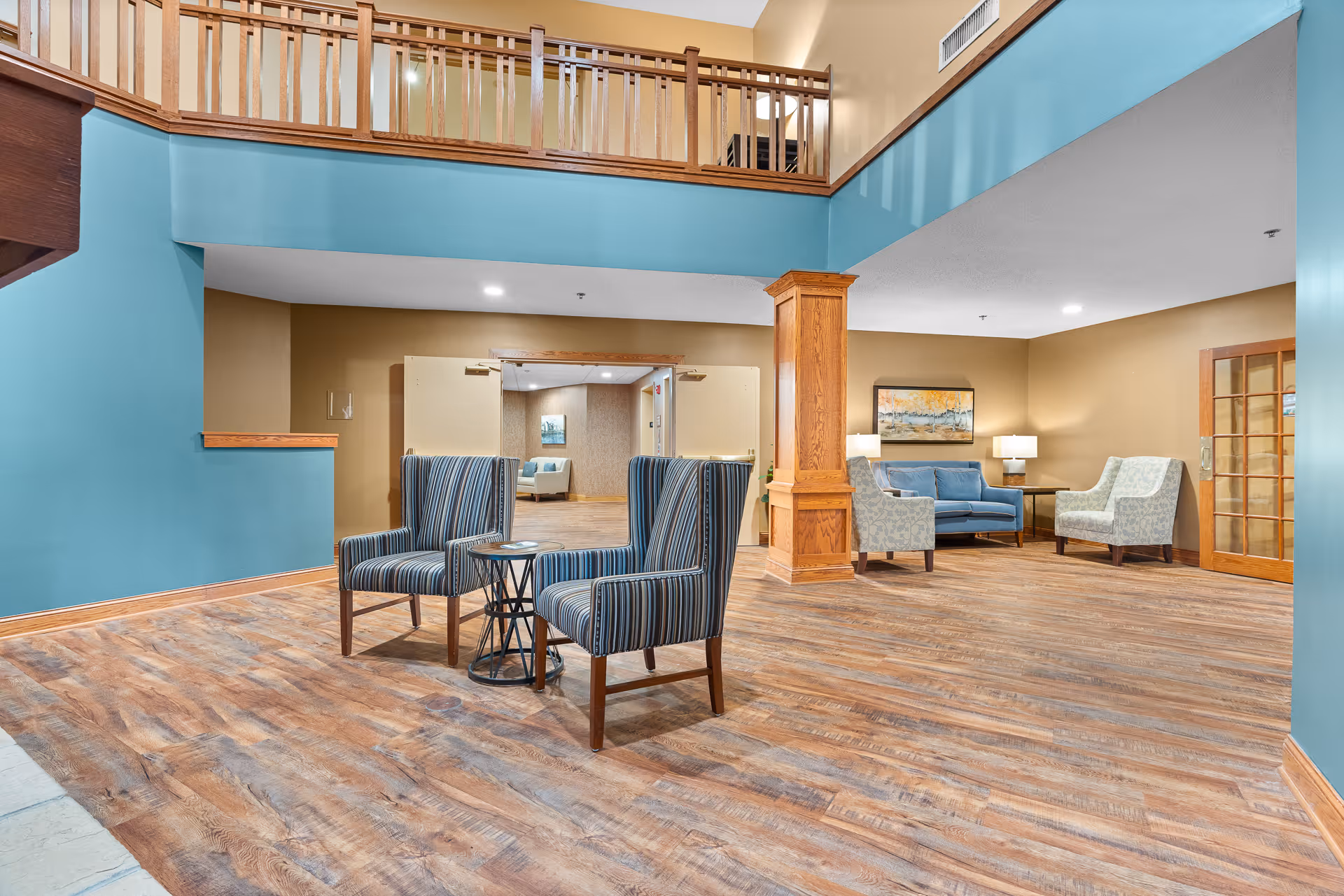A spacious and well-lit common area with wooden flooring and a high ceiling. The room features two striped armchairs with a small round table between them in the foreground. In the background, there is a seating area with a blue sofa, two patterned armchairs, a side table with a lamp, and a framed artwork on the wall. The walls are painted in a combination of blue and beige tones, and there is a wooden railing on the upper level overlooking the space.