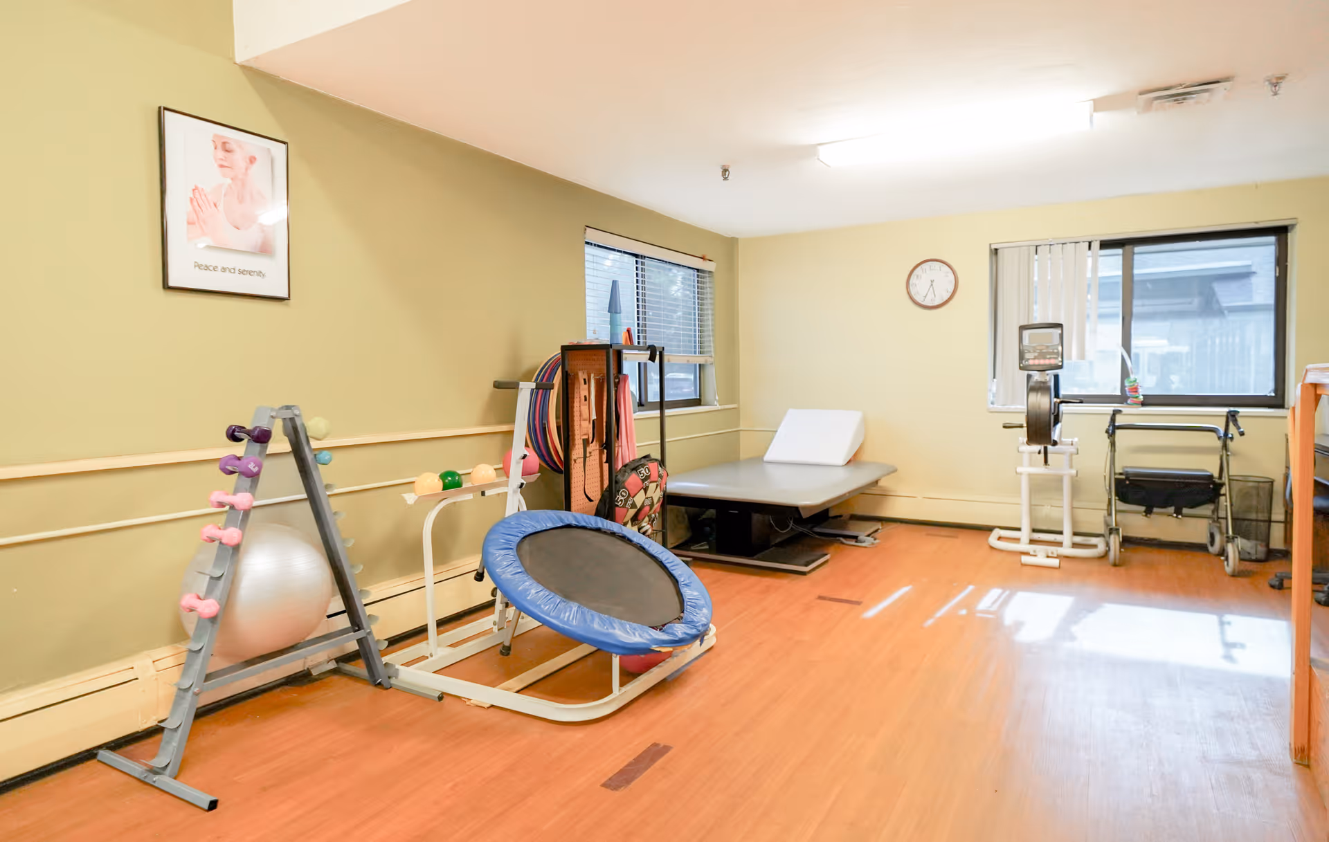 A small exercise room with wooden flooring and light yellow walls. The room contains various fitness equipment including a rack of small dumbbells, a silver exercise ball, a mini trampoline, resistance bands, a padded exercise table, a rowing machine, and a walker. There are two windows with blinds, a wall clock, and a framed picture on the wall that reads 'Peace and serenity'.