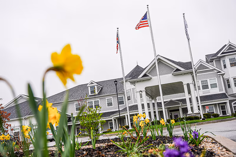 Front entrance of a multi-story senior living building with flagpoles and daffodils in the foreground.