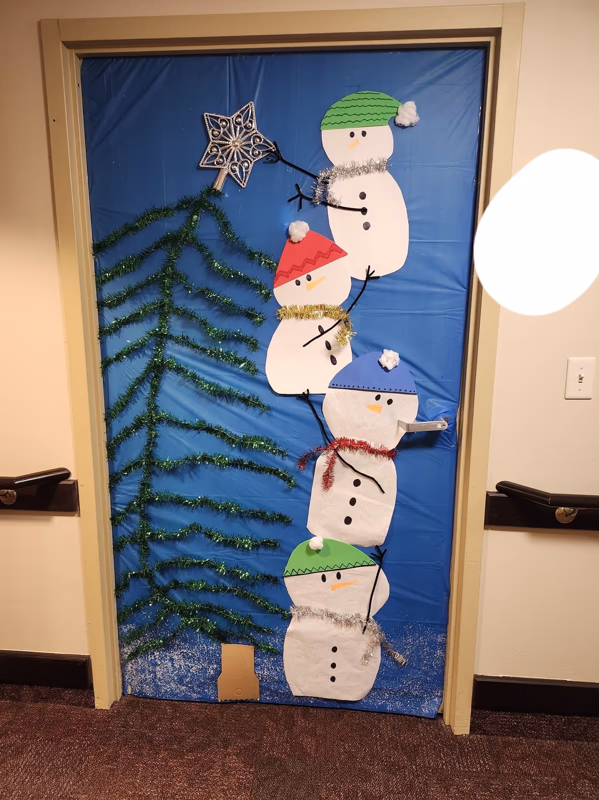 A hallway door decorated with blue paper, tinsel shaped like a tree, and stacked paper snowmen wearing colorful hats and garlands.
