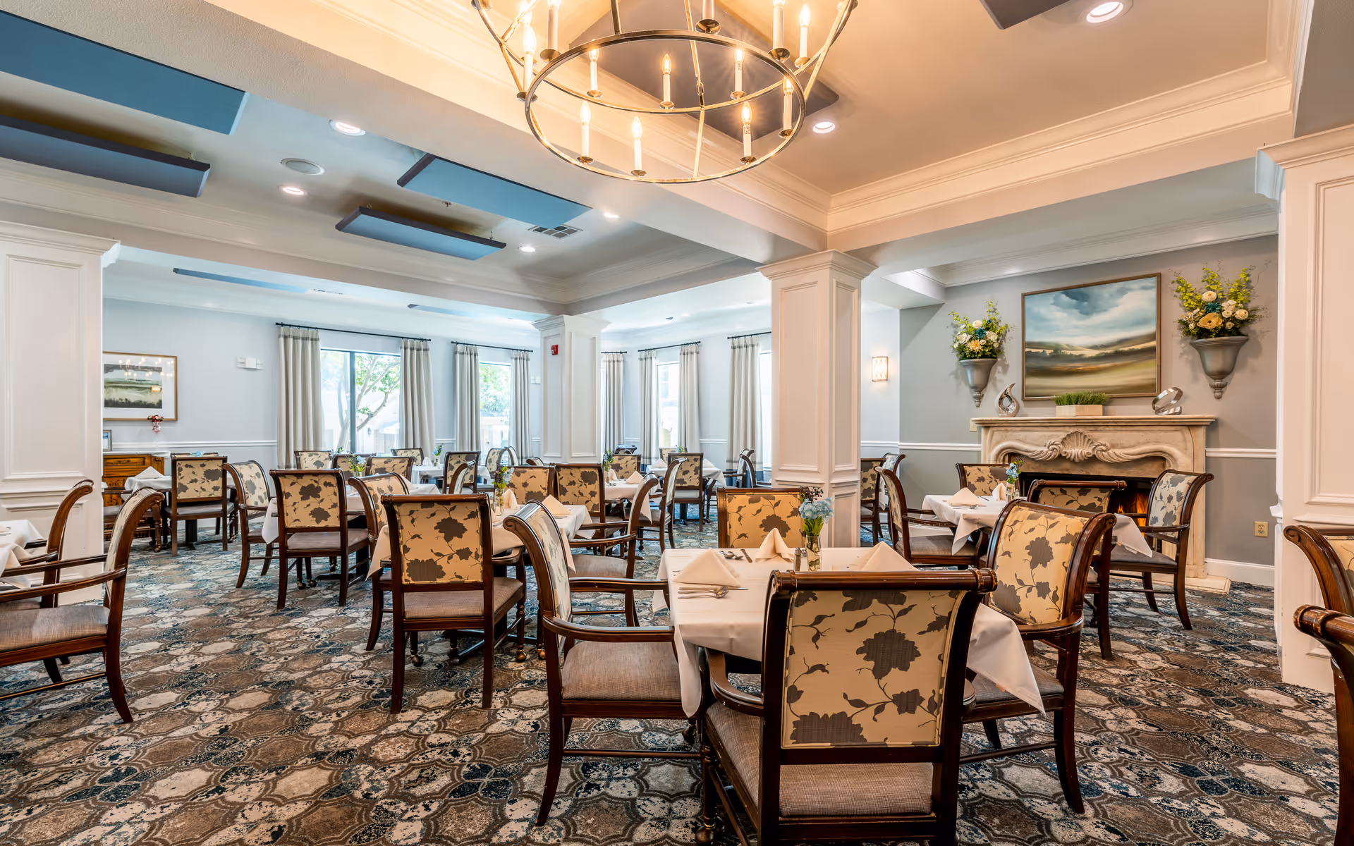A spacious, elegantly decorated dining room with multiple tables covered in white tablecloths and set with napkins. The chairs have floral patterned upholstery. The room features large windows with curtains, a decorative fireplace with floral arrangements and a landscape painting above it, and a chandelier hanging from the ceiling.