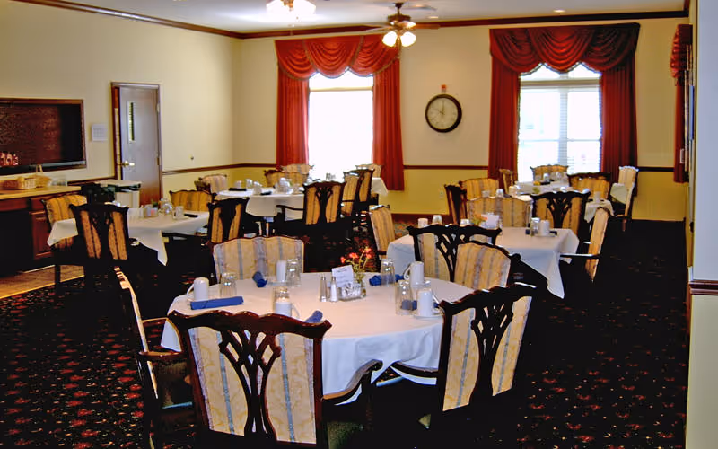 A dining room with several round tables covered with white tablecloths, each set with cups, glasses, and napkins. The chairs have wooden backs with intricate designs and cushioned seats. The room has red curtains on the windows, a wall clock, and ceiling fans with lights.