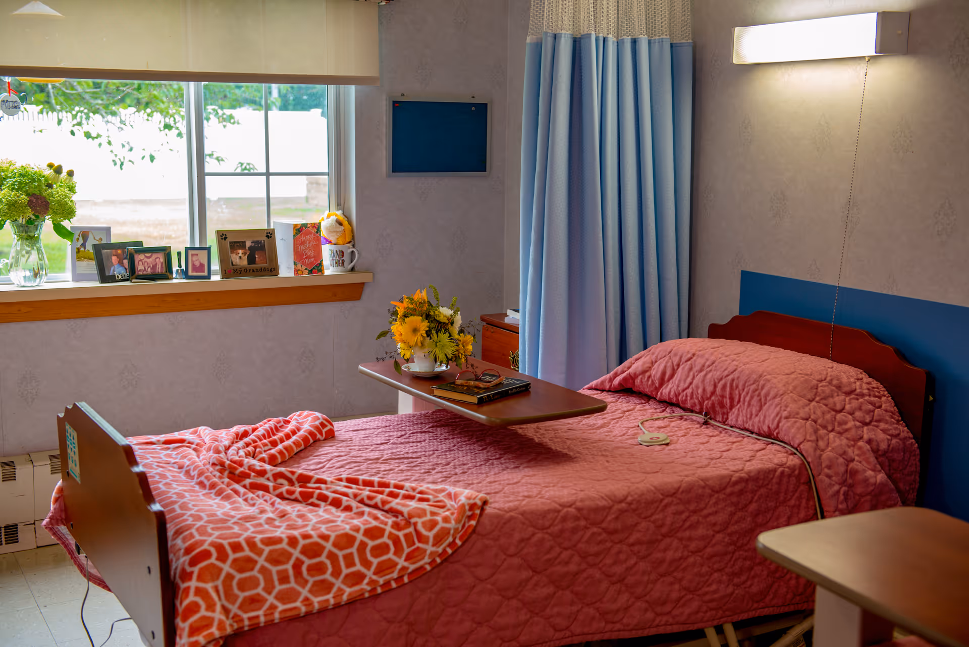 A cozy bedroom in a senior living facility with a single bed covered in a pink quilt and an orange patterned blanket. A wooden overbed table holds a vase of yellow flowers and some books. The room has a window with a windowsill displaying framed photos and a small plant. Light blue curtains hang beside the bed, and a wall-mounted light fixture illuminates the room.
