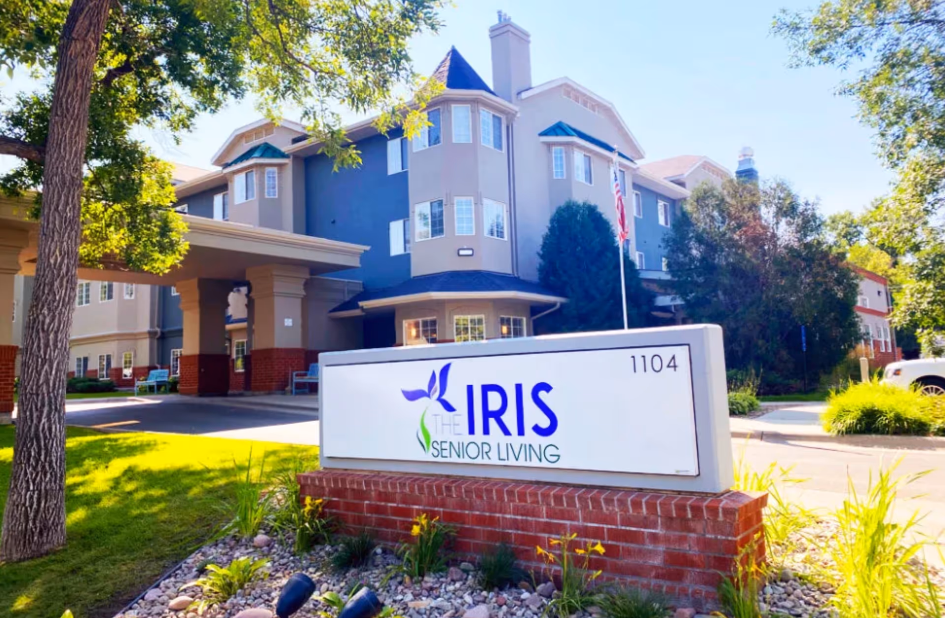 Exterior view of The Iris Senior Living facility showing a multi-story building with beige and blue walls, surrounded by trees and greenery. In the foreground, there is a large sign with the facility's name and address number 1104, set on a brick base with decorative plants and rocks around it.
