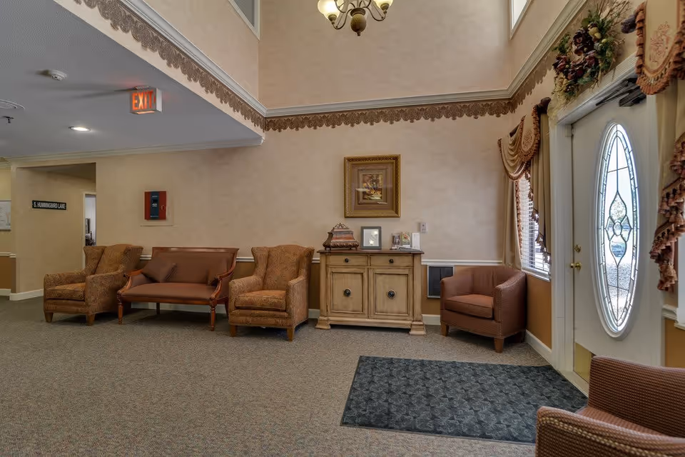 A cozy seating area in a senior living facility with two patterned armchairs, a wooden loveseat with cushions, a small wooden cabinet with decorative items and framed pictures, a window with ornate curtains, and a glass-paneled door with an oval design. The walls are beige with a decorative border near the ceiling, and there is a carpeted floor with a dark mat near the door.