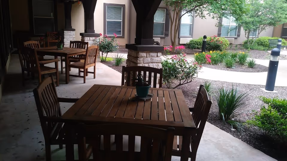 Outdoor covered patio area with wooden tables and chairs, potted plants on the tables, and a landscaped garden with bushes, flowers, and small trees in the background near building windows.