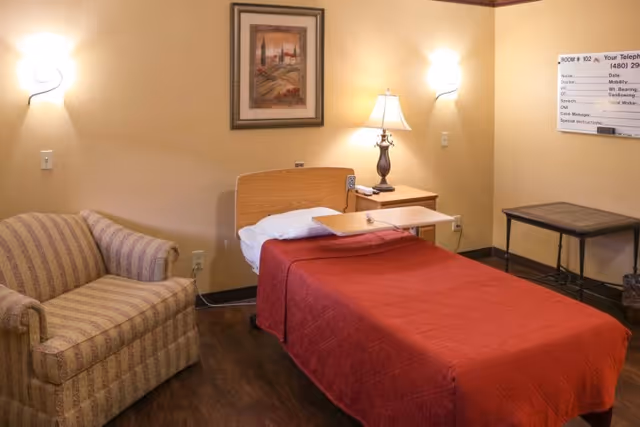 A cozy room with a single bed covered in a red quilt, a wooden headboard, and an overbed table. Next to the bed is a nightstand with a decorative lamp. There is a striped armchair to the left and a framed painting on the wall above the bed. The room has warm lighting from two wall sconces and a lamp, with a small table and a whiteboard on the right side.