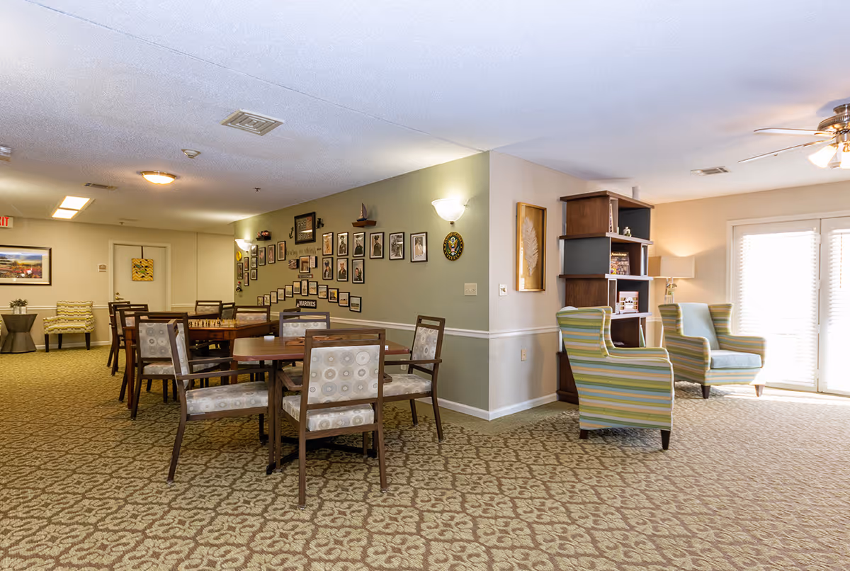 Well-lit common room with several dining tables and chairs, striped armchairs by a bookshelf, and framed photos on a wall.