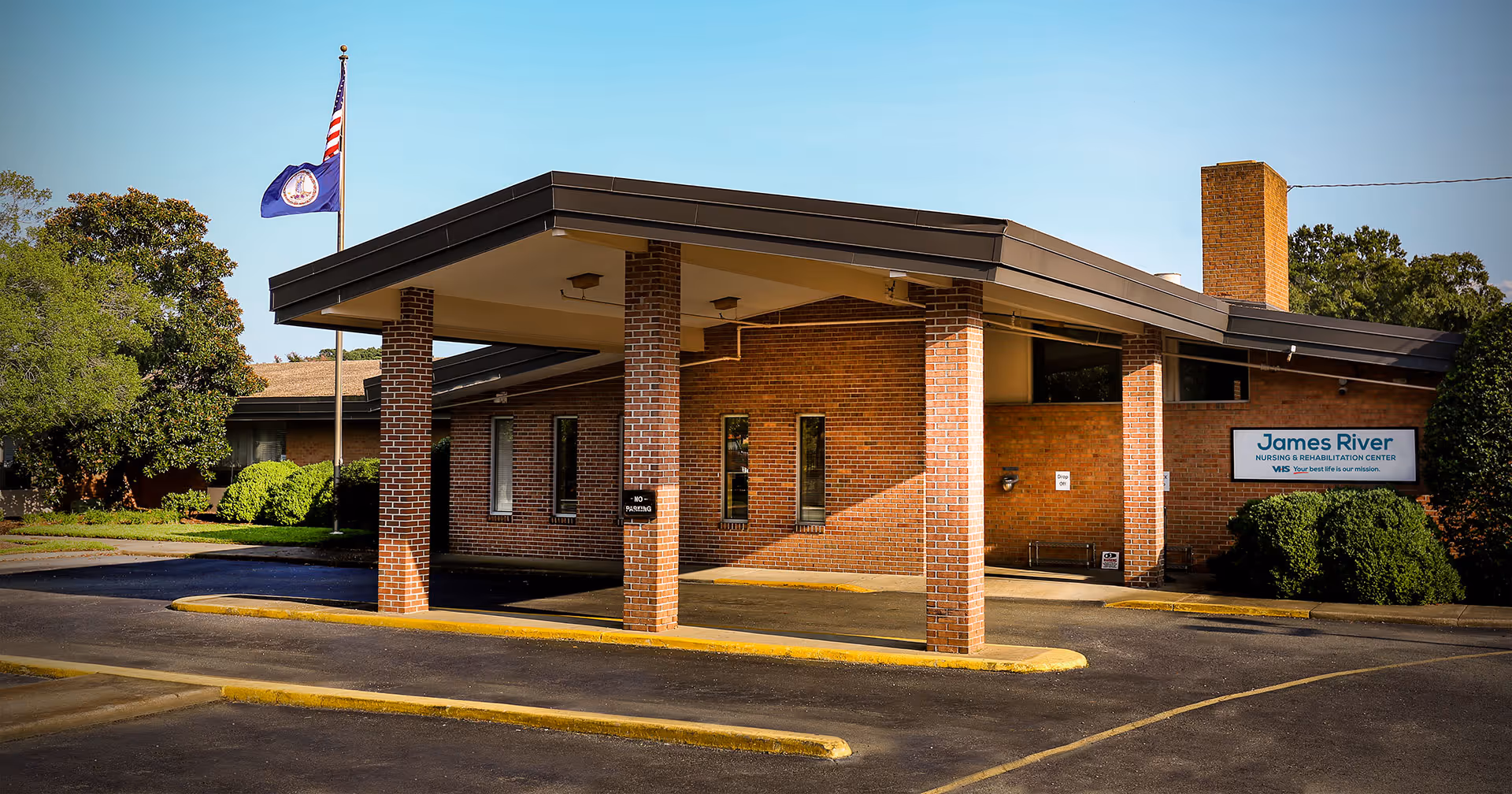 Exterior front view of James River Nursing & Rehabilitation Center building with a covered entrance supported by brick columns. There are two flags on flagpoles to the left, trees and bushes surrounding the building, and a clear blue sky.