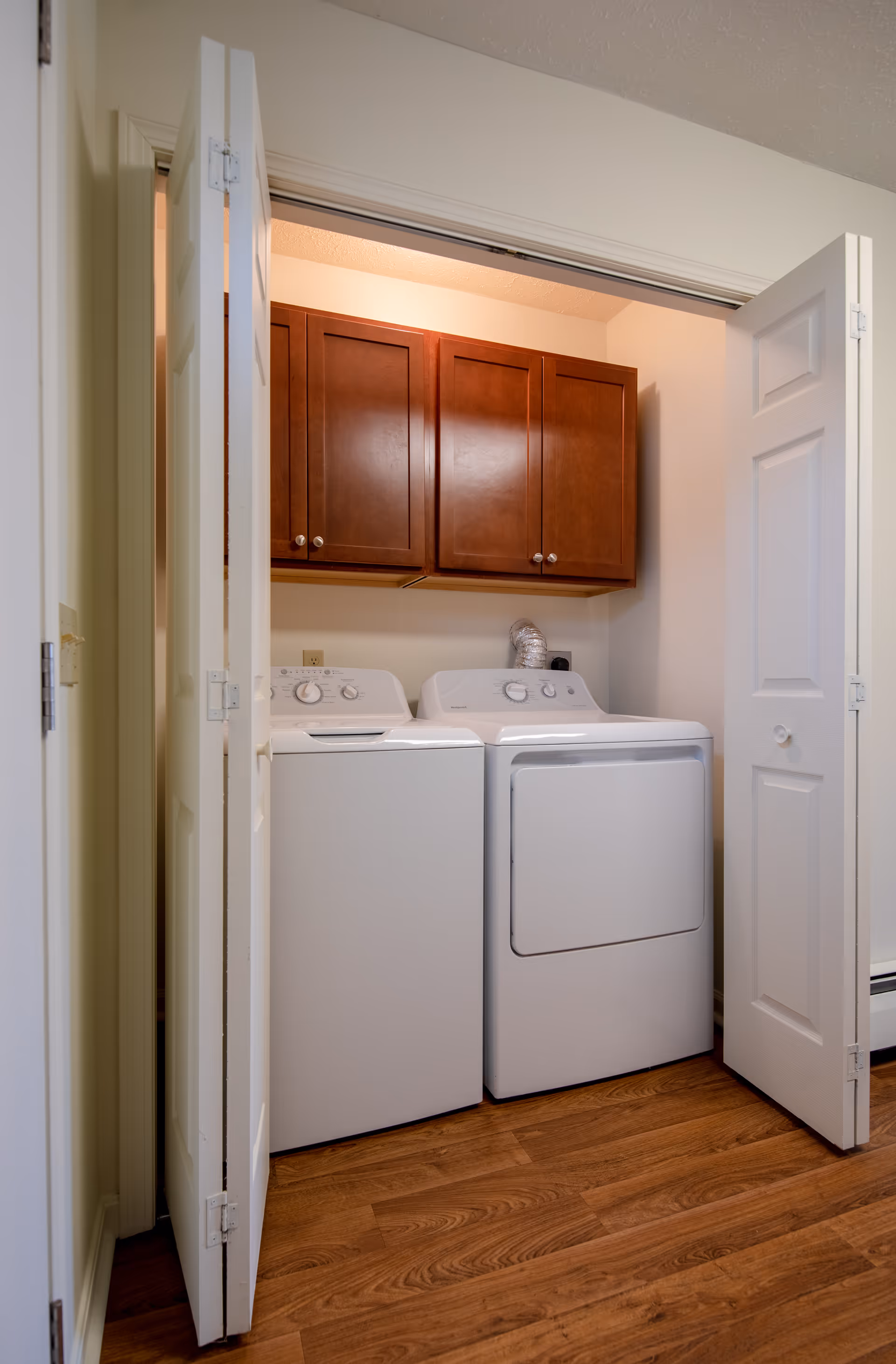 Laundry area with a white washing machine and dryer side by side, enclosed by bi-fold white doors. Above the machines are wooden cabinets with a dark brown finish. The floor is a medium-tone wood.