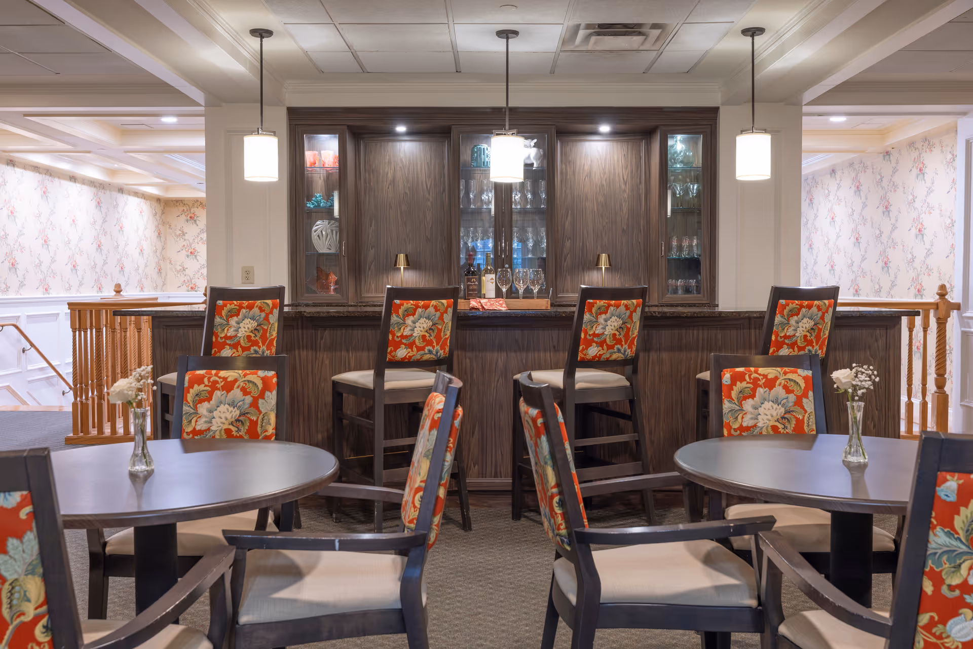 Interior view of a common area with a bar counter and several round tables with chairs. The chairs have floral patterned upholstery. Behind the bar counter are wooden cabinets with glass doors displaying glassware. The room is softly lit with pendant lights hanging from the ceiling and floral wallpaper on the walls.