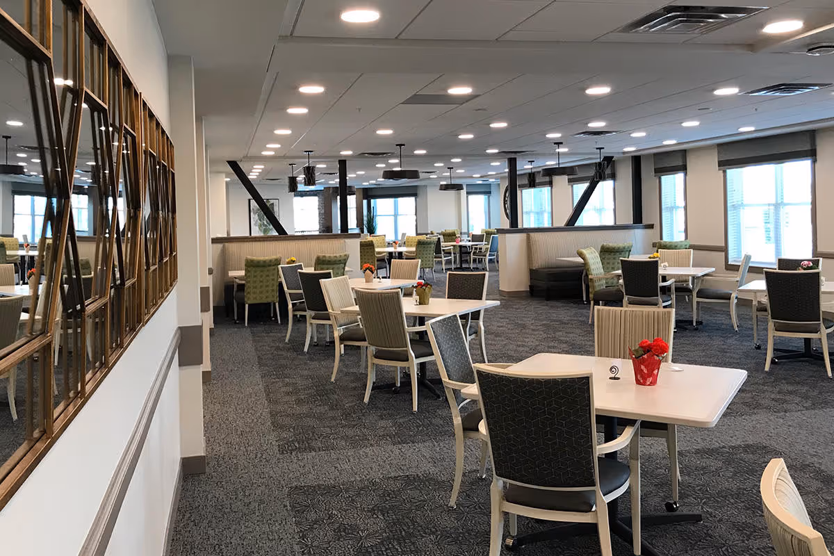 A spacious dining room with multiple tables and chairs arranged neatly. The room has large windows allowing natural light to fill the space, and decorative mirrors on one wall. Some tables have small flower arrangements as centerpieces.