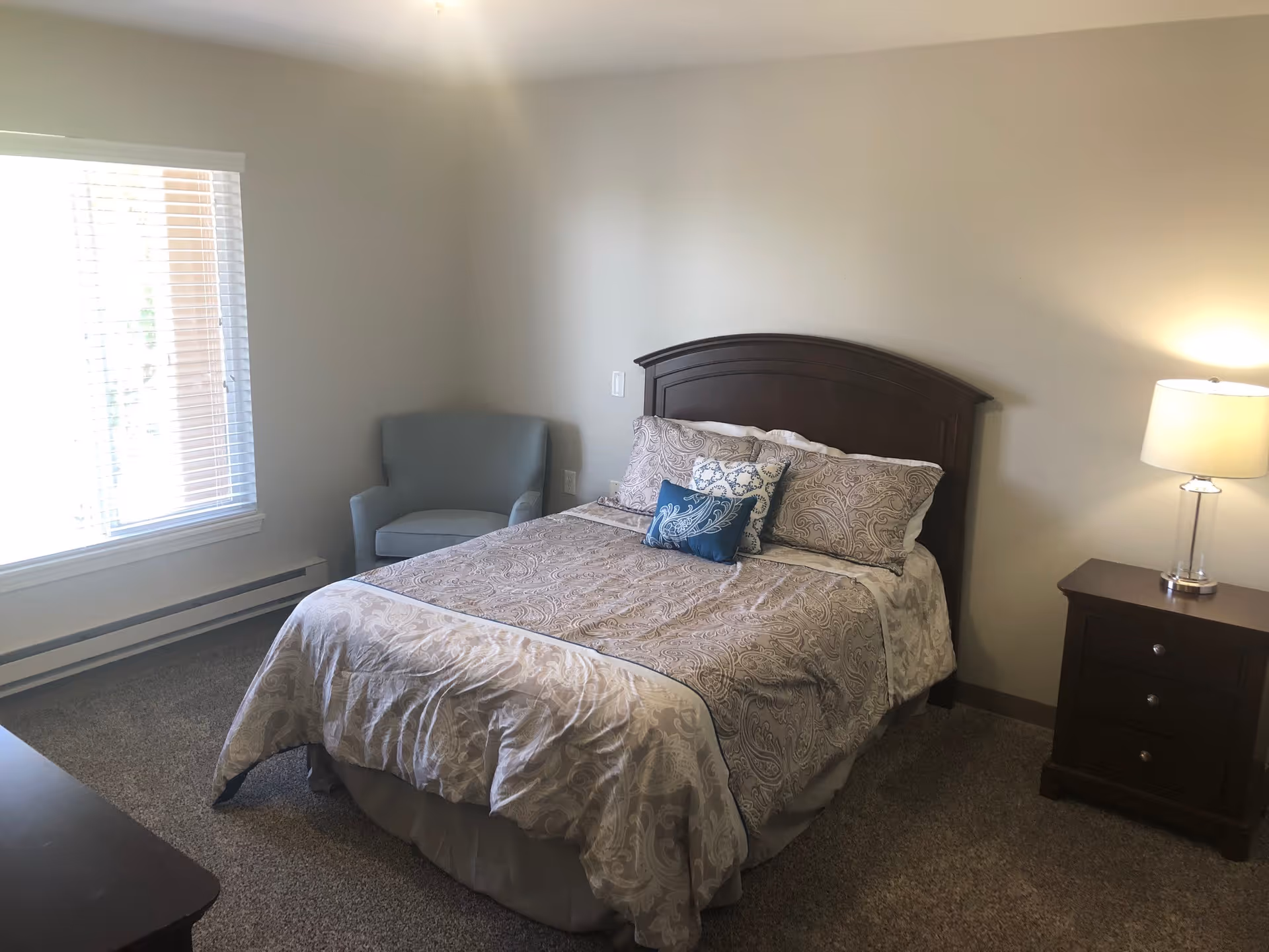 A bedroom with a bed featuring a dark wooden headboard, beige patterned bedding, and three decorative pillows. To the right of the bed is a dark wooden nightstand with a lamp on it. To the left of the bed is a light blue armchair near a window with white blinds. The room has beige walls and carpeted floor.