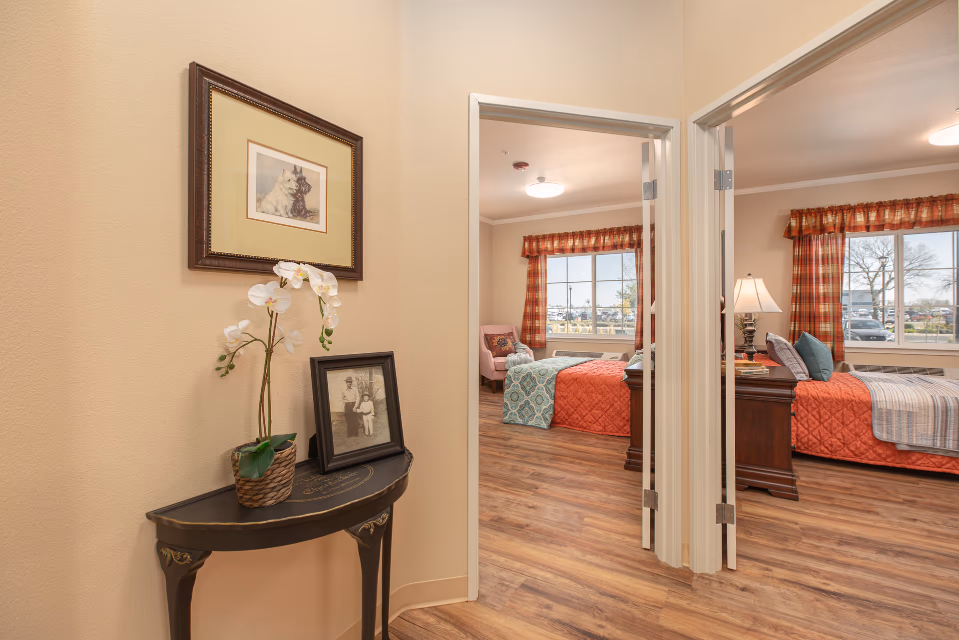View of a hallway corner with a small decorative table holding a potted orchid and a framed black-and-white photo. On the wall above the table is a framed picture of two dogs. The hallway leads to two bedrooms, each with a bed covered in colorful quilts, a window with red plaid curtains, and wooden flooring throughout.