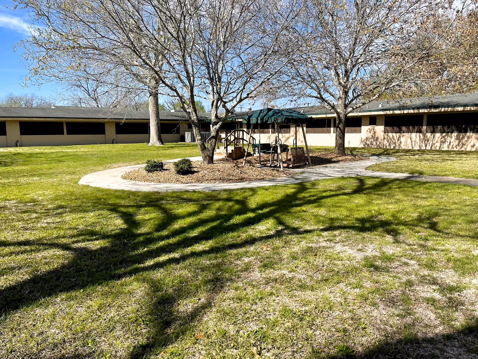 Outdoor courtyard area with green grass, two leafless trees, a circular paved walkway, and a shaded swing bench. The background shows a single-story building with multiple windows under a clear blue sky.