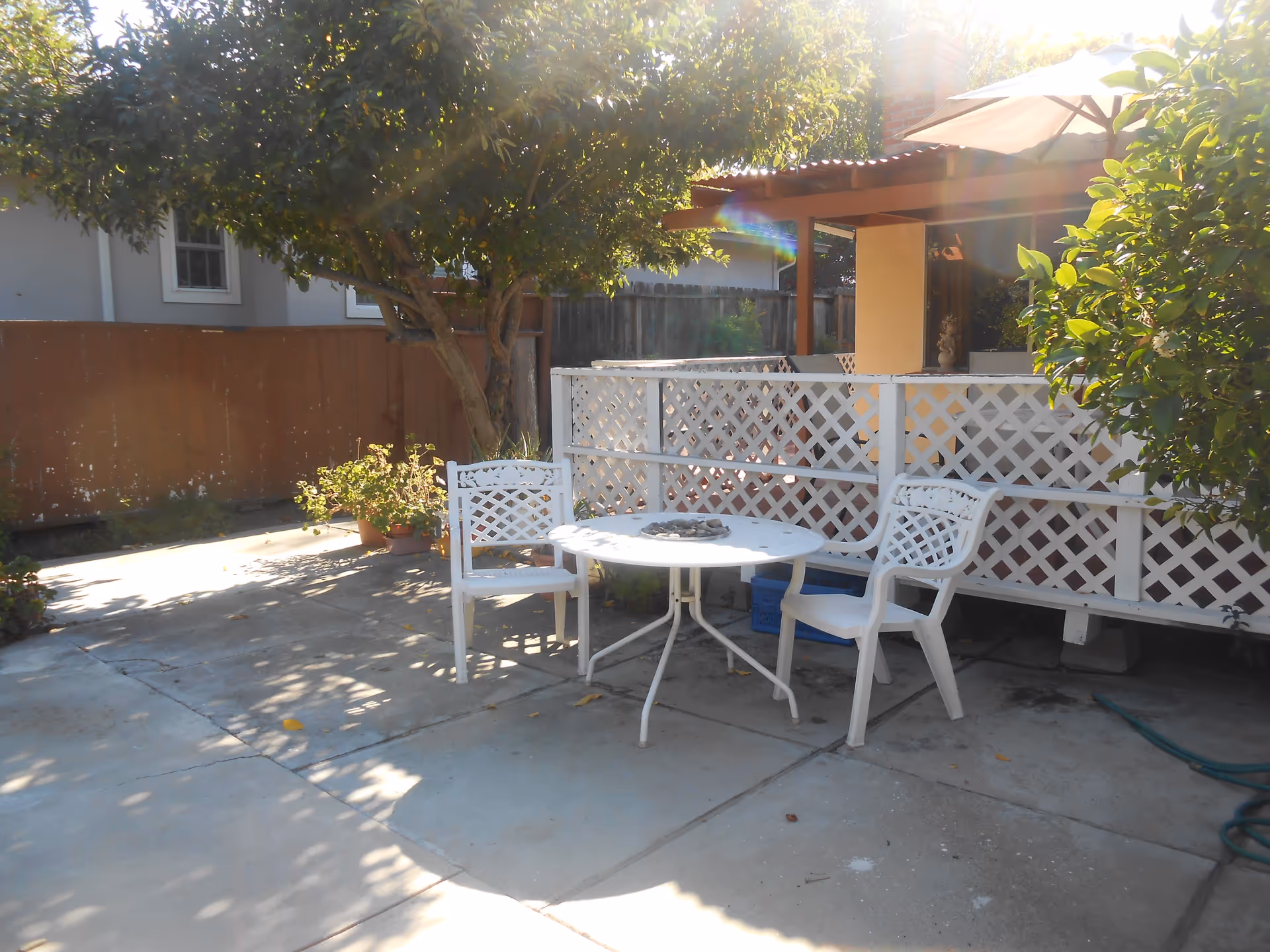 Sunlit outdoor patio with a white plastic table and chairs beside a lattice fence and trees.