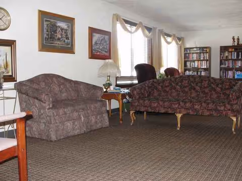Cozy senior living room featuring patterned couches, a lamp, windows, and a tall bookshelf.