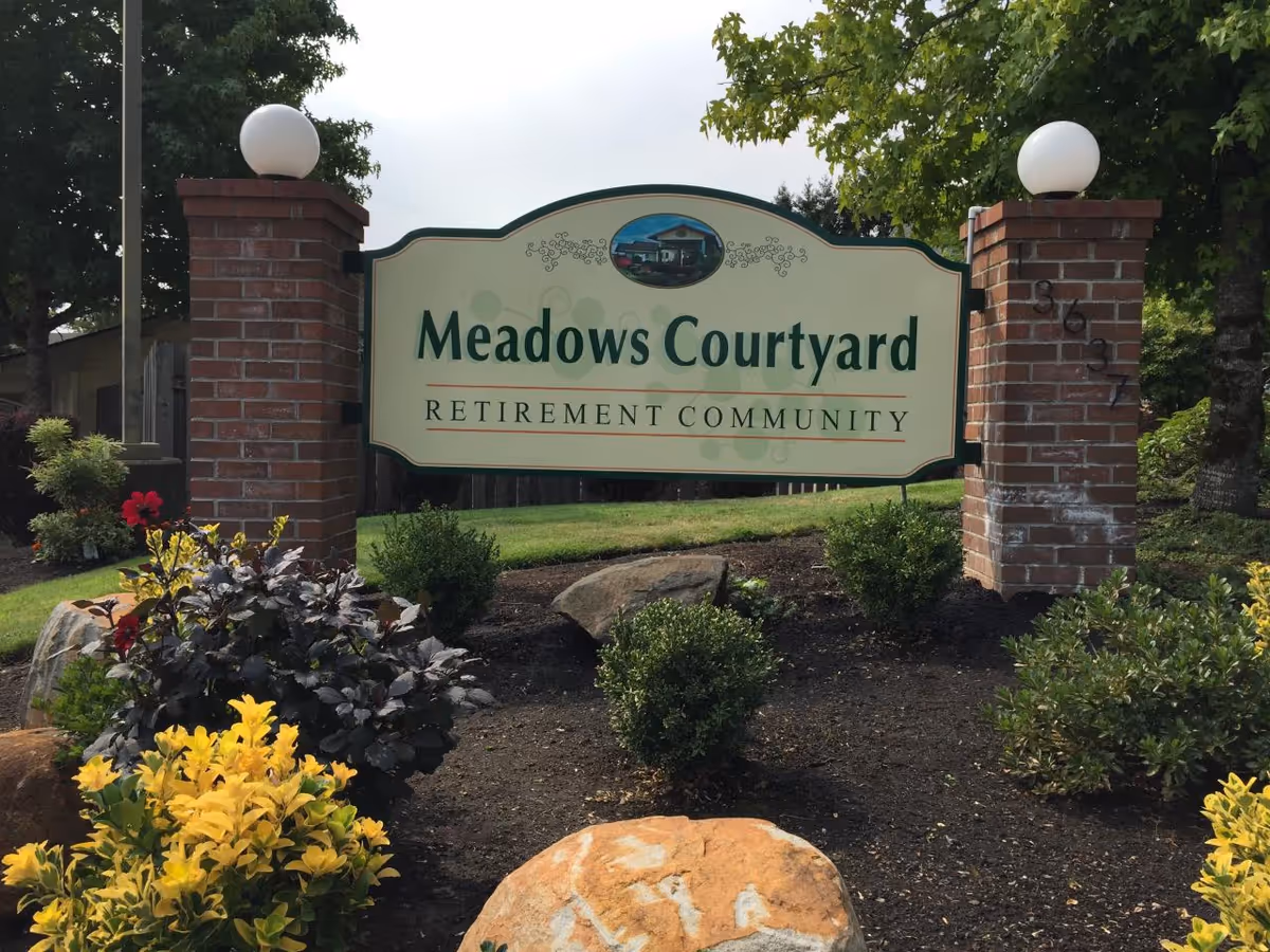 Entrance sign reading "Meadows Courtyard Retirement Community" mounted between two brick pillars with landscaping in front.