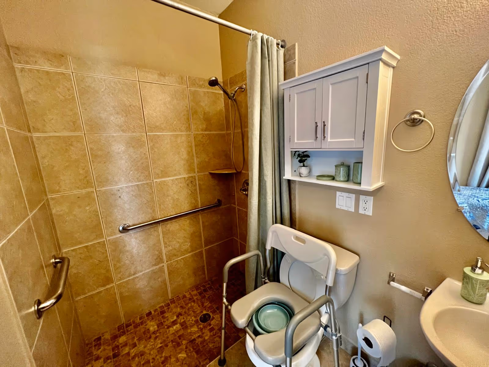 Bathroom with a tiled walk-in shower equipped with grab bars and a shower curtain. There is a white cabinet mounted on the wall above a toilet, which has a raised toilet seat with armrests and a green basin placed on it. A round mirror, a towel ring, a sink with a soap dispenser, and a toilet paper holder are also visible.