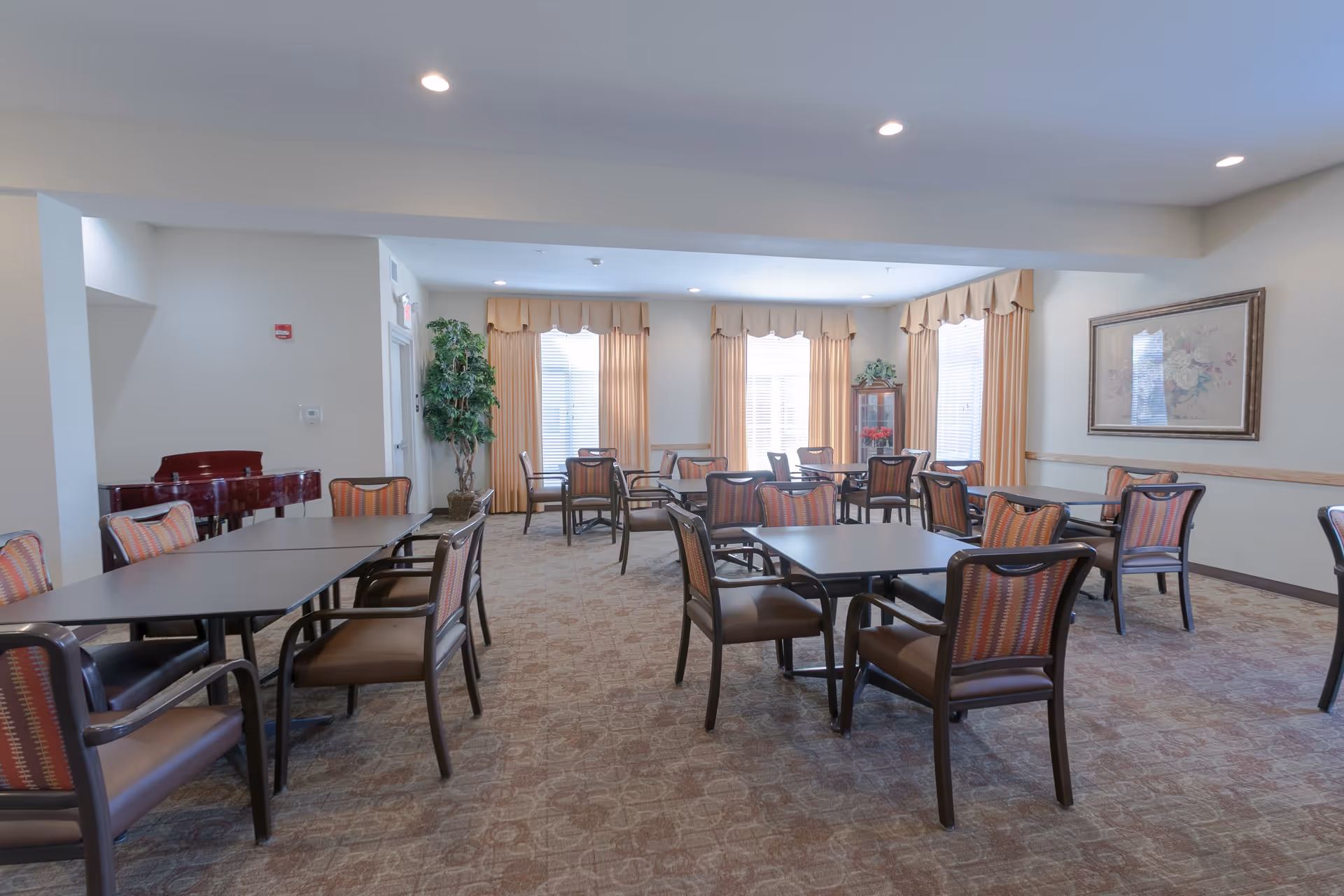 Spacious common dining room with multiple tables and chairs, large windows with curtains, a piano, and framed artwork.
