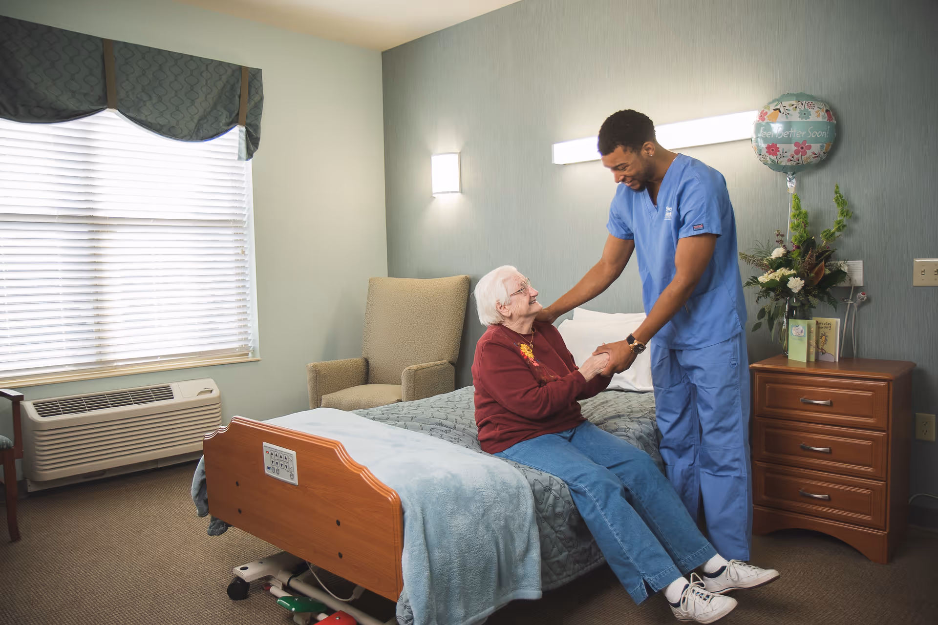 An elderly woman sitting on a bed in a nursing facility room, smiling and holding hands with a male caregiver dressed in blue scrubs who is standing beside her. The room has a window with blinds, a beige armchair, a wooden nightstand with flowers, cards, and a 'Get Well Soon' balloon.