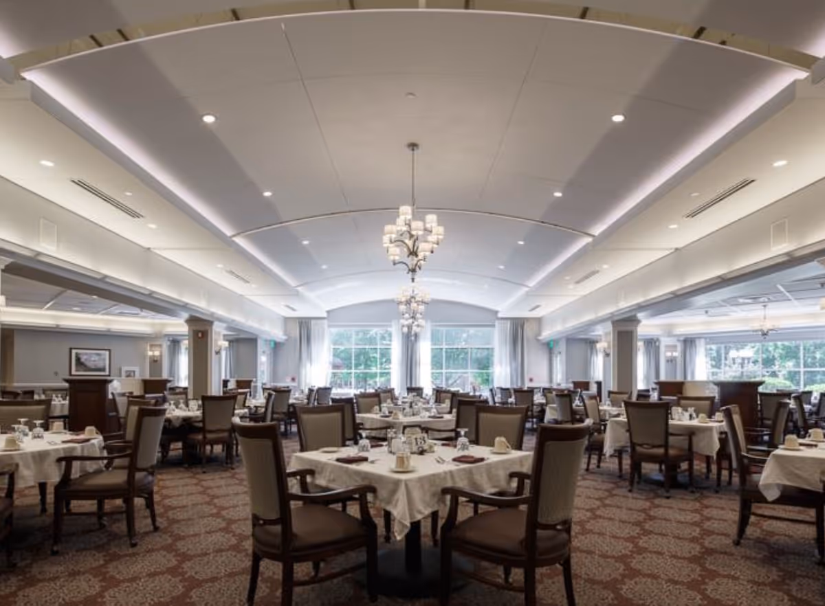 Large elegant dining room with multiple set tables, upholstered chairs, chandeliers, and large windows.