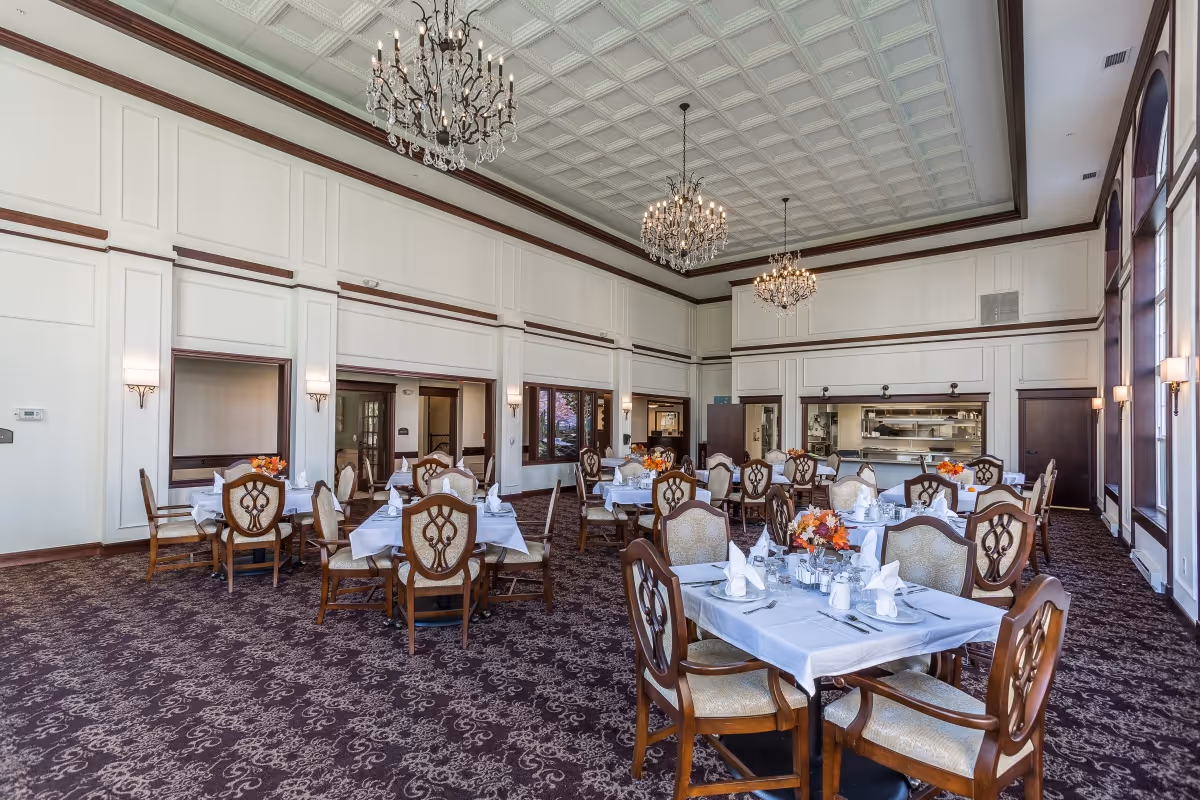 Elegant dining room with multiple tables set with white tablecloths, napkins, and floral centerpieces. The room features high ceilings with ornate chandeliers, large windows allowing natural light, and patterned carpet flooring. The walls are paneled in white with dark wood trim.