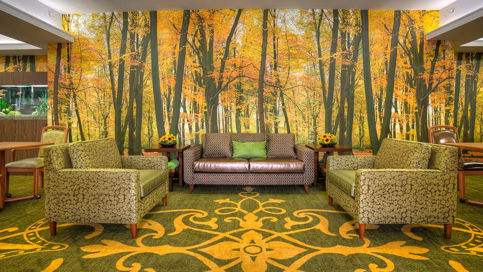 A cozy living room area with a green and yellow patterned carpet, two green patterned armchairs, and a brown patterned sofa with green cushions. The walls feature a large mural of a forest with trees in autumn colors. There are small wooden side tables with flower arrangements and a fish tank visible on the left side.