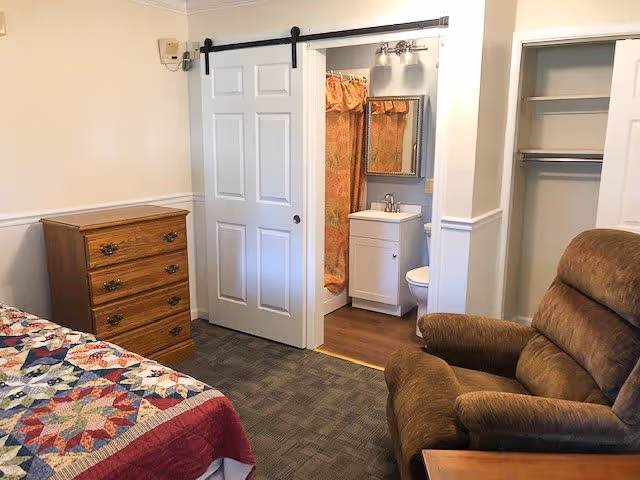 A cozy senior living bedroom featuring a bed with a colorful quilt, a wooden chest of drawers, a brown recliner chair, an open closet with shelves and hanging rod, and a sliding barn door leading to a small bathroom with a sink, mirror, toilet, and shower with a patterned curtain.