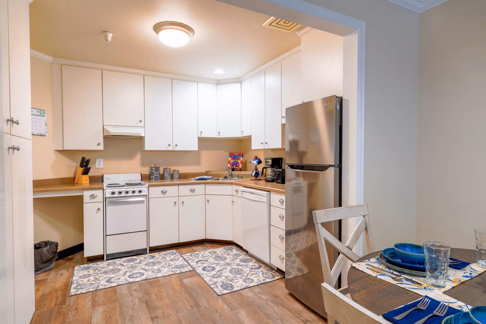 A clean and well-lit kitchen with white cabinets and beige countertops. The kitchen features a small white stove, a stainless steel refrigerator, a dishwasher, and a coffee maker. There are two patterned rugs on the wooden floor. To the right, part of a dining table is visible with blue plates, glasses, and silverware set on a floral placemat.