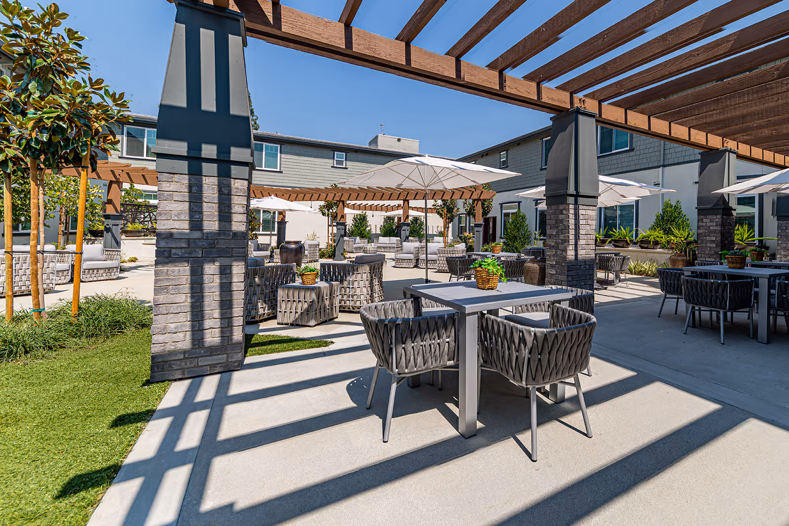 Outdoor patio area at Allara Senior Living with multiple tables and chairs under large umbrellas, surrounded by greenery and shaded by wooden pergolas attached to brick pillars, with a clear blue sky overhead.
